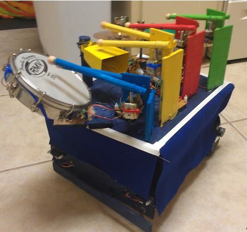 The assembled DrumBot on the floor: a blue-skirted mobile base carrying the drum kit on its upper deck, with the snare cantilevered off the left side and the colour-coded sticks poised over each drum