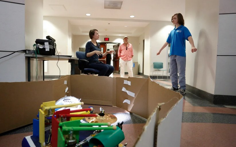 Demo day in a University of Florida engineering hallway: DrumBot's colour-coded drum kit in the foreground inside a cardboard arena, with me seated behind it explaining the robot while spectators look on