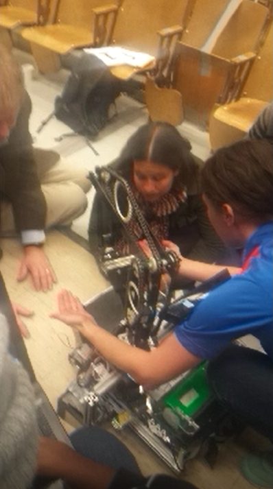 Teammates crouched on the floor around the built arm, working on its chain drive and linkage between runs