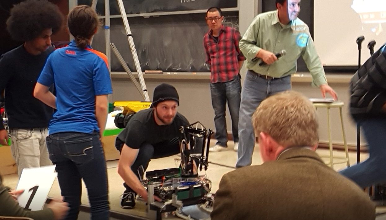 The robot at the competition, a teammate crouched over it making a final adjustment while judges and the other team look on
