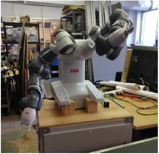 The ABB YuMi robot standing on wooden blocks on a workbench in the lab, both arms raised, with lab equipment and shelving behind it