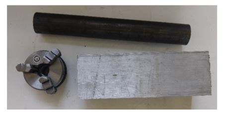 The stock for the spindle laid out: a length of steel round bar, a rectangular aluminium block for the housing, and the mini lathe three-jaw chuck