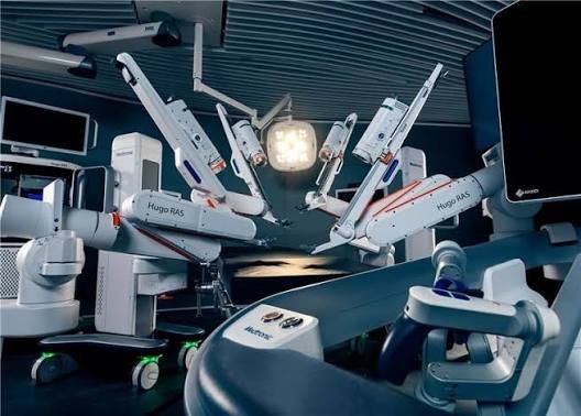 The Hugo robotic-assisted surgery system: several white robotic arms on mobile carts arranged around an operating table under surgical lights, with the surgeon's open console in the foreground