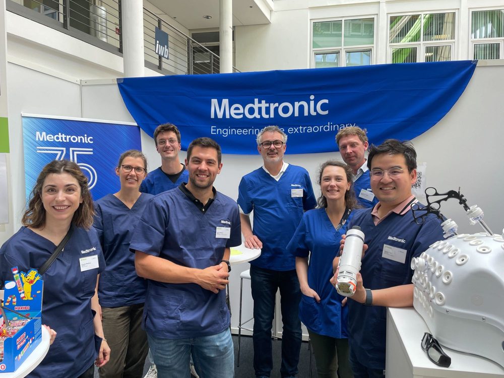 Nine colleagues in Medtronic scrubs under a Medtronic banner at a university atrium booth, one holding a device beside a model head