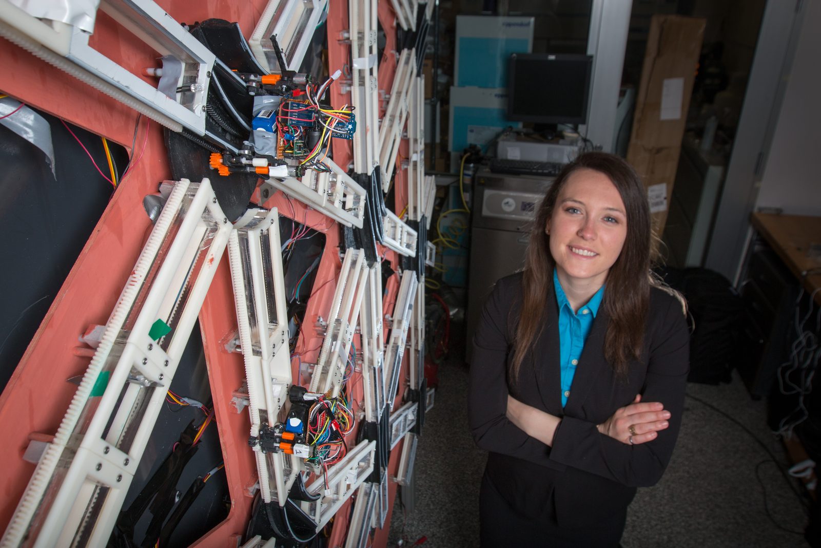 Standing beside the Multi-Track Elevator prototype in the d'Arbeloff Laboratory — a wall-mounted triangular grid of white rails with rack teeth, carriages and control electronics mounted across it