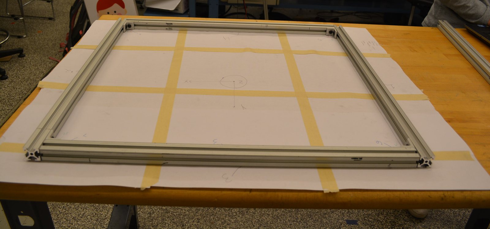 The 80/20 aluminium frame I built, laid out on a bench over paper marked with a taped grid and hand-drawn coordinate axes, with corner gussets bolted at each corner