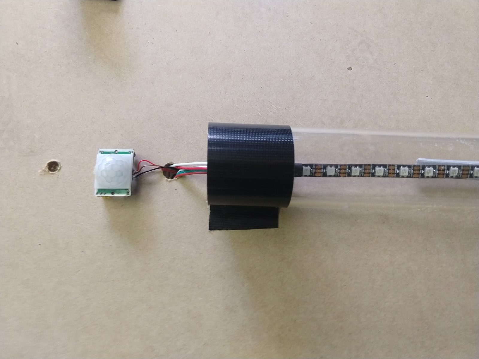 A Parallax passive-infrared motion sensor taped to a panel next to an acrylic tube with an LED strip run through it — the mock UV lamp assembly before installation