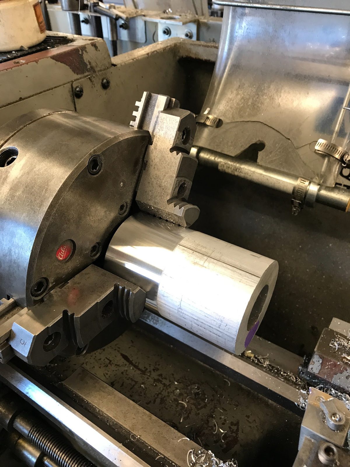 A stepped aluminium bar held in a three-jaw chuck on a manual lathe, its outside diameter turned and its bore opened: the spindle housing blank