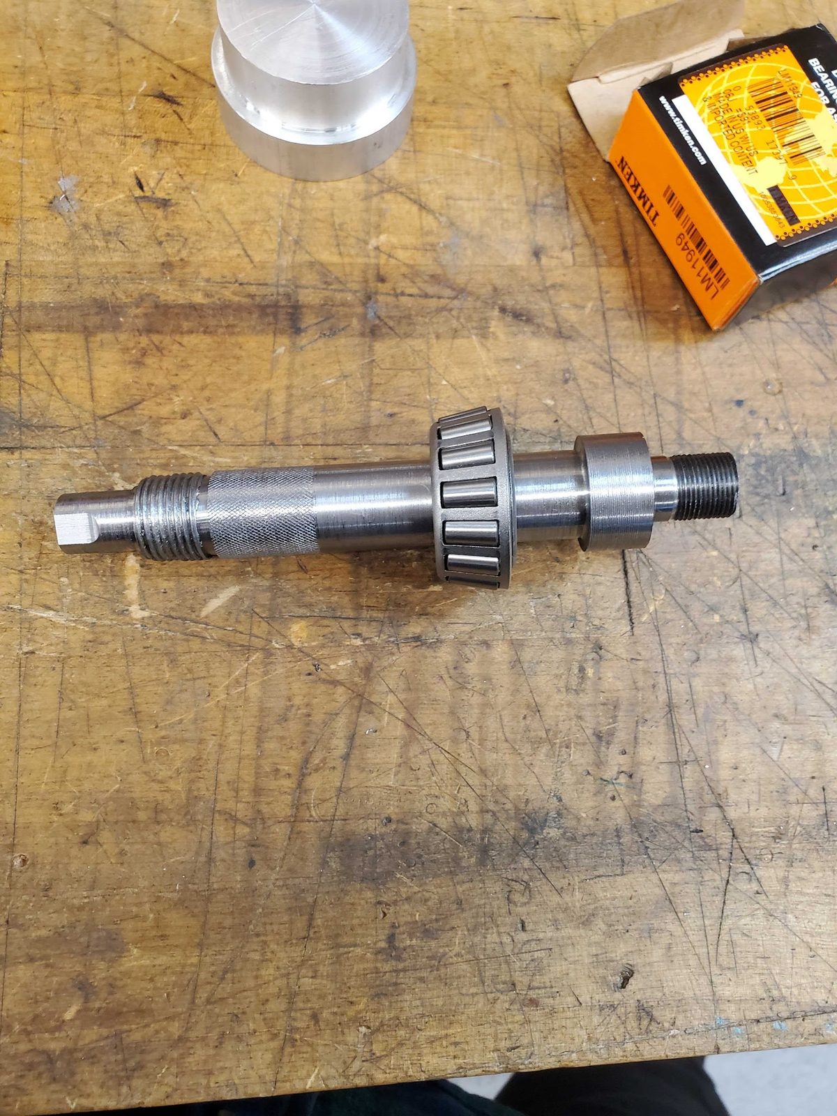 The machined steel shaft on a workbench with a Timken LM11949 tapered roller bearing cone pressed onto its front seat, the bearing's box beside it
