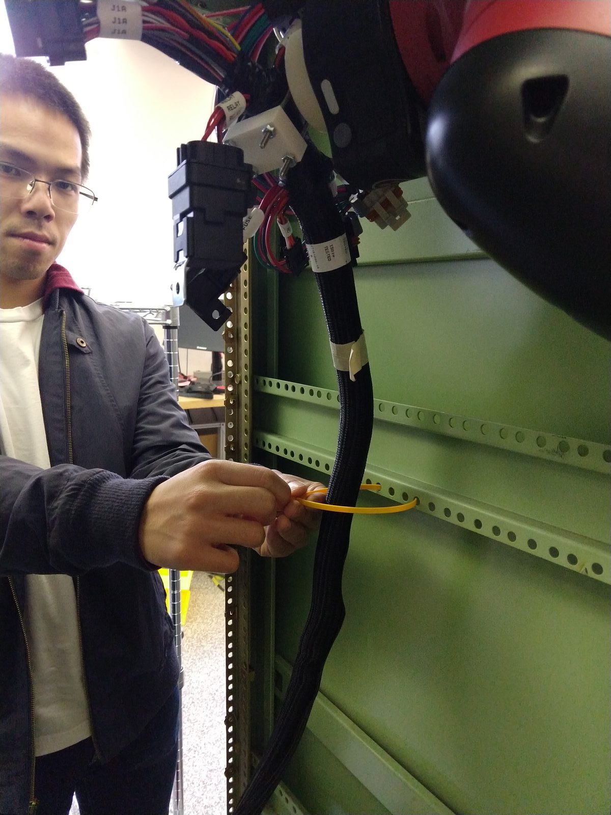 A worker's hands securing the harness with a yellow zip tie while a Sawyer gripper holds it against the cabinet rail