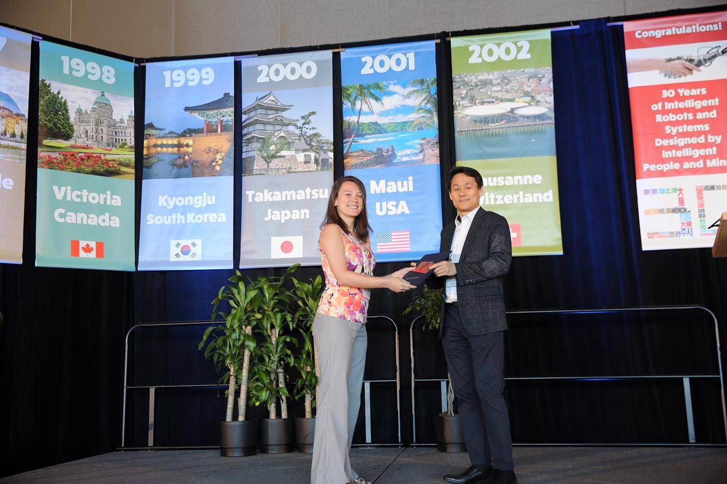 Me on stage at IROS 2017 in Vancouver, accepting the Best Application Paper Award certificate in front of the conference's banners of past host cities